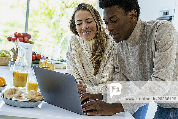 Adult man typing on laptop while leaning on kitchen counter with girlfriend