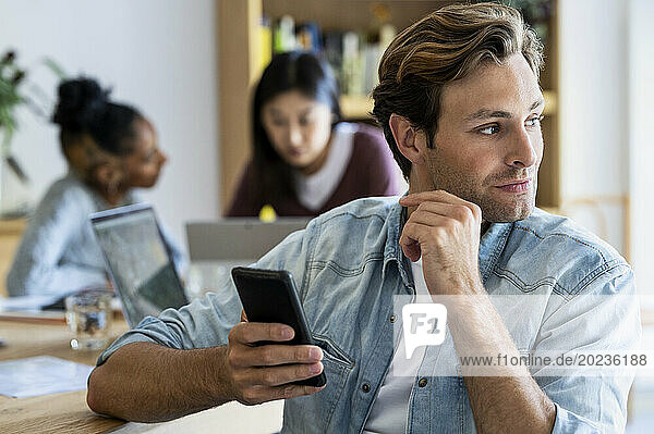 Young adult entrepreneur holding smart phone with coworkers on the background