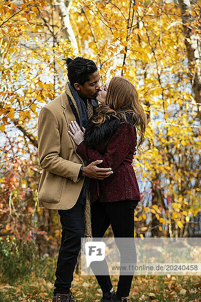 A mixed race couple standing by the shore of a lake hugging and kissing each other while spending quality time together during a fall family outing in a city park; Edmonton  Alberta  Canada