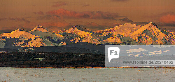 Snow-covered mountain range glowing with the warm light of sunrise and warm glowing clouds with foothills and snow-covered field in the foreground  West of Calgary  Alberta; Alberta  Canada