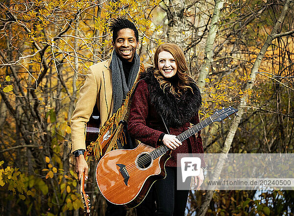 Portrait of a mixed race married couple holding acoustic guitars and smiling at the camera while spending quality time together during a fall family outing in a city park; Edmonton  Alberta  Canada