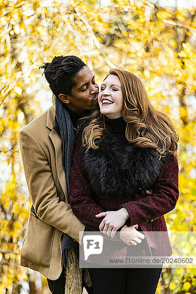 A mixed race married couple hugging with husband kissing wife on the cheek while spending quality time together during a fall family outing in a city park; Edmonton  Alberta  Canada