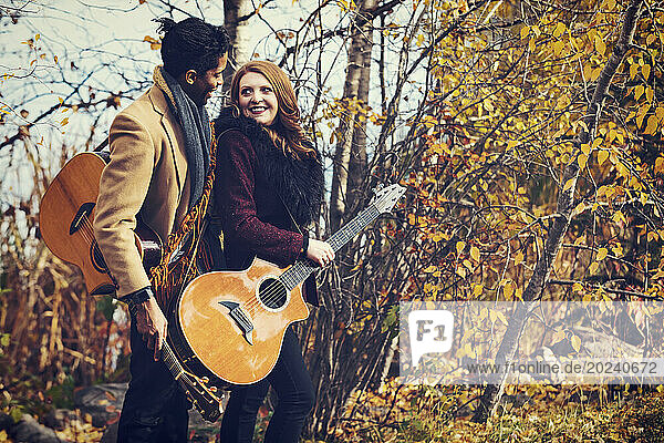Portrait of a mixed race married couple holding acoustic guitars and smiling at each other  spending quality time together during a fall family outing in a city park; Edmonton  Alberta  Canada