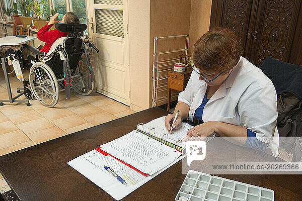 HAD (Hospitalization At Home). Follow-up of an elderly patient at her home by a caregiver. Nursing assistant filling out the transmission book with the elderly and disabled patient installed in her wheelchair in the background.