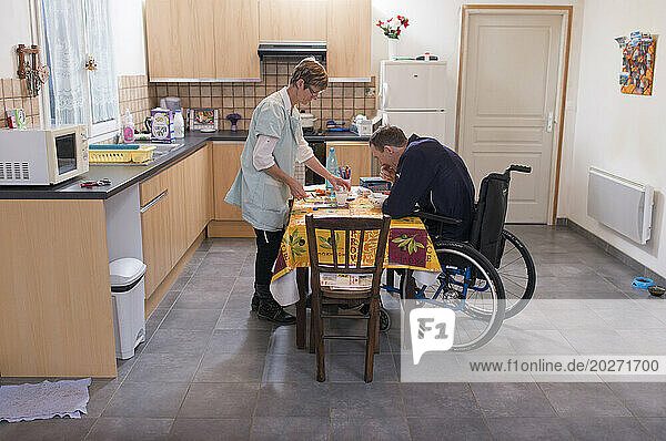 ADMR 62 - Home Help in Rural Areas  Pas de Calais. Muriel  Auxiliary of Social Life (AVS) works at the home of Mr. T. who is severely handicapped by Multiple Sclerosis. Muriel must help her with all the daily tasks. Carer helping a disabled person in a wheelchair during his breakfast.