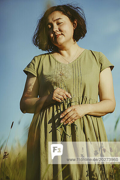 Mature woman holding dandelion and standing in field on sunny day