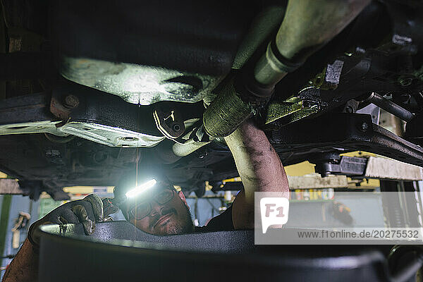 Mechanic using flashlight and examining parts below vehicle at workshop