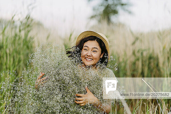 Cheerful woman wearing hat and holding bunch of gypsophila flowers in field