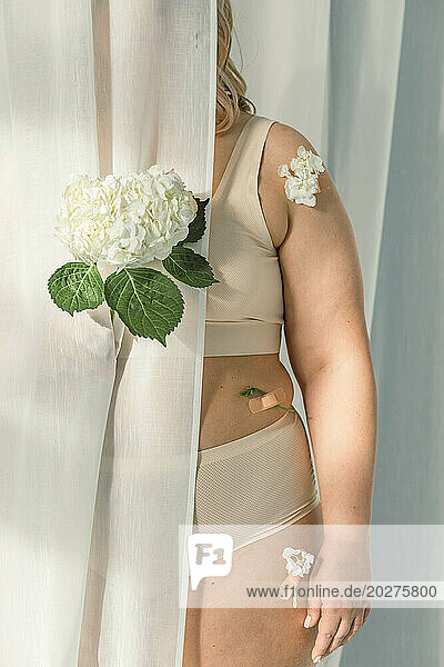 Woman with flowers on body hiding behind white curtain