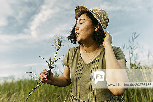 Playful woman wearing hat and blowing on dandelion in field