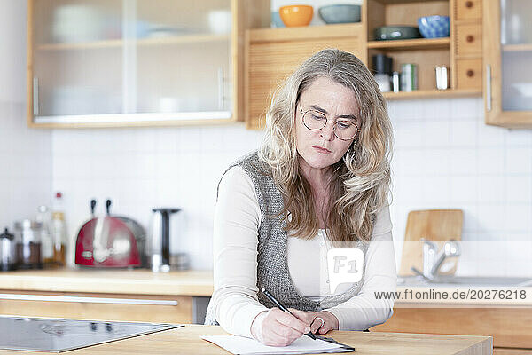 Serious mature woman writing on paper in kitchen at home