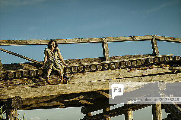 Carefree woman sitting on wooden bridge under blue sky