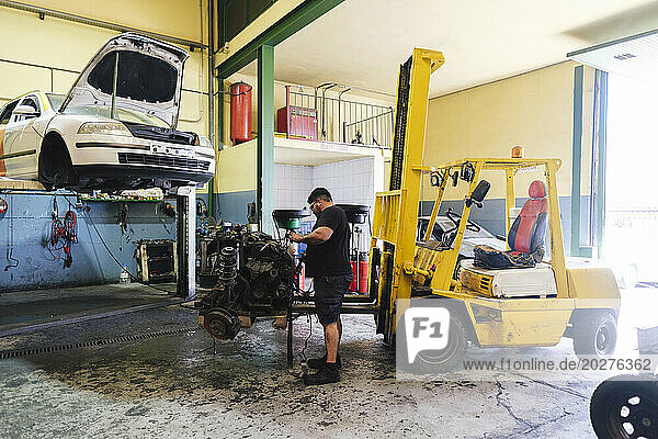 Auto mechanic dismantling engine parts near forklift at workshop