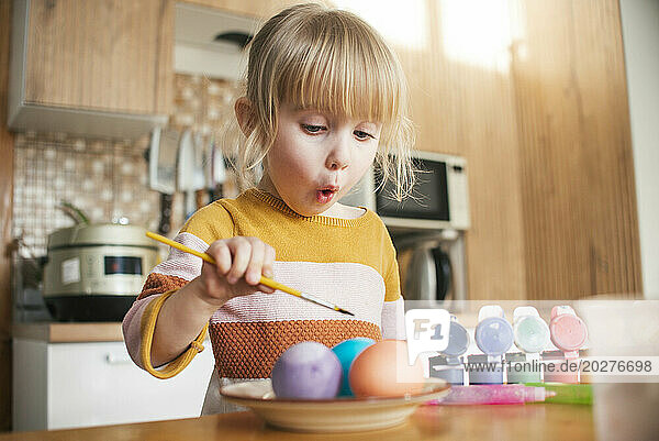 Surprised girl coloring Easter eggs with paintbrush at home