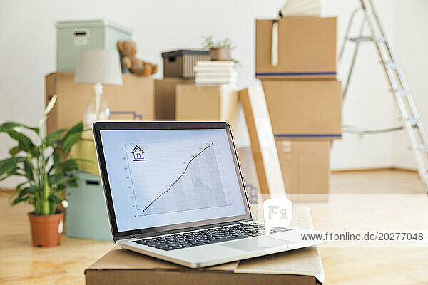 Rising line graph on laptop screen in front of cardboard boxes in an empty room in a new home