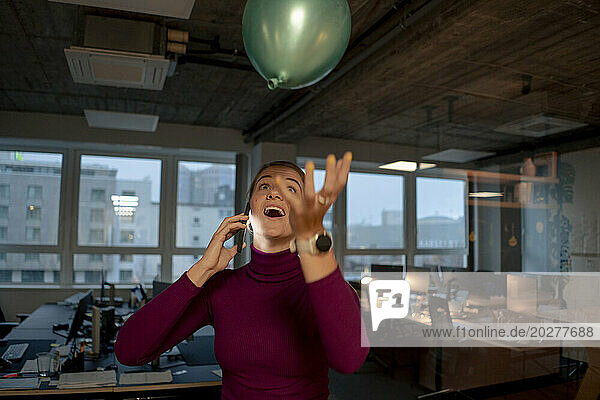 Playful businesswoman talking on smart phone and catching balloon in office