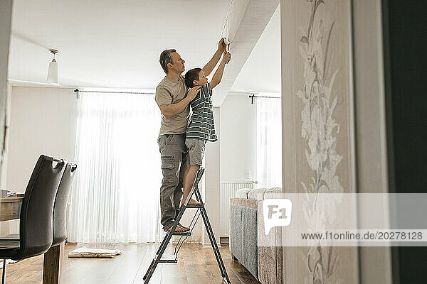 Son and father peeling old wallpaper standing on step ladder at home