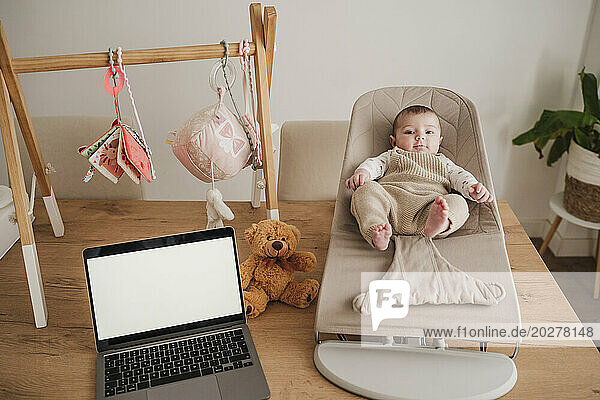 Cute baby girl sitting on bouncer chair near laptop at home
