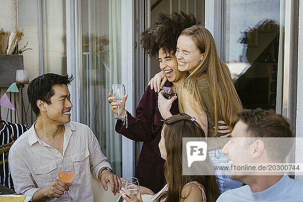 Cheerful woman embracing female friend and enjoying drinks while celebrating dinner party in balcony