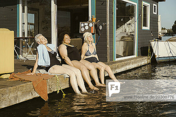 Smiling senior female friends dipping legs in water while relaxing during vacation on houseboat