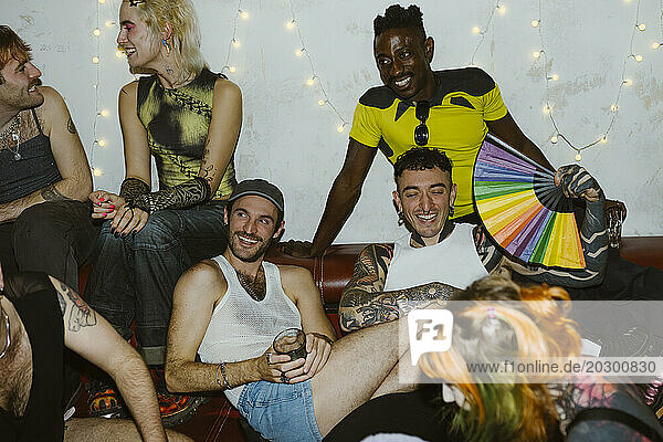 Happy non-binary friends sitting on sofa at enjoying party at illuminated nightclub