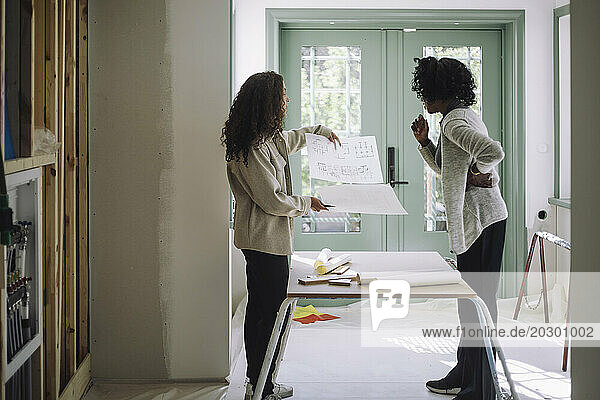 Side view of client and female architect comparing blueprints while standing near table at under construction apartment