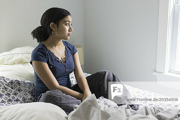Biracial teen girl sits on un unmade bed looking depressed