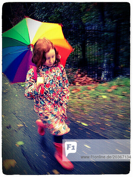 A young redheaded girl hurries to school on a rainy day wearing a floral rain coat red boots and carrying a rainbow umbrella -