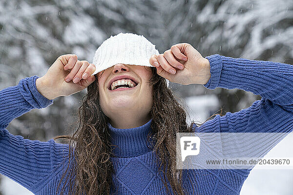 Playful woman hiding face with knit hat in winter forest