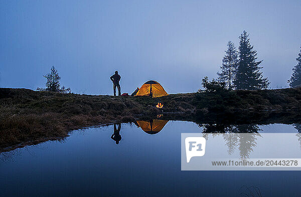 Senior man standing near tent and camping at lake shore