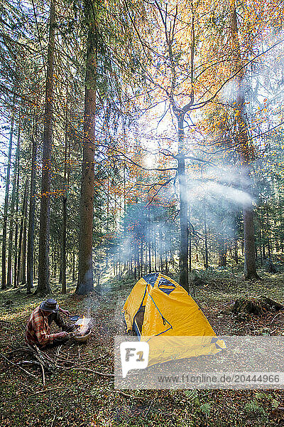 Senior man cooking food near yellow tent in forest