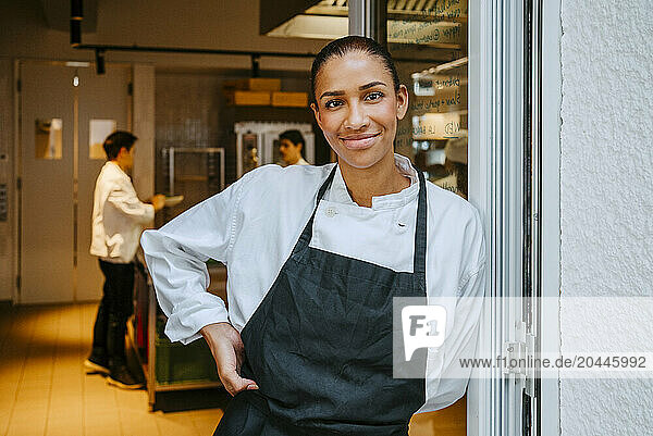 Portrait of happy female chef leaning on door at commercial kitchen