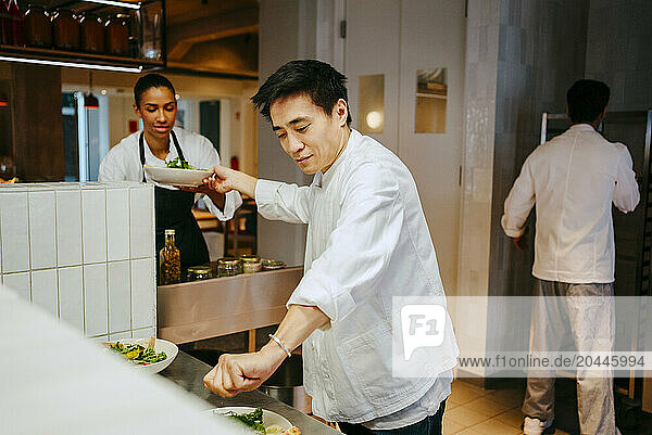 Male chef passing salad bowl to female coworker while finishing salad at commercial kitchen