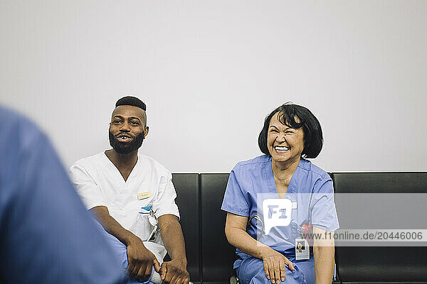 Happy male and female medical professionals discussing while sitting on chair near wall in hospital