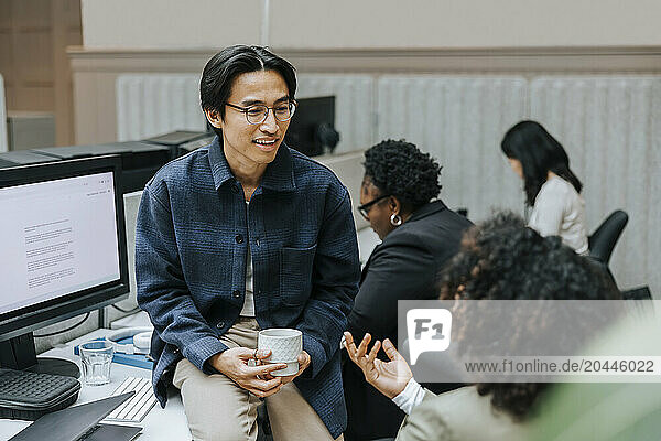 Businesswoman holding coffee cup while sitting on desk and discussing with female colleague at office