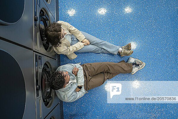 Young couple sitting together leaning on washing machines in laundromat