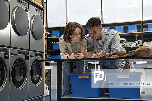 Young man and woman using smart phones leaning on table at laundromat