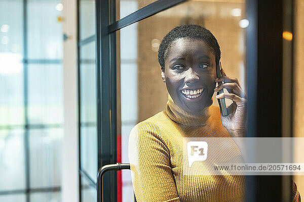 Happy businesswoman talking through smart phone leaning on glass wall