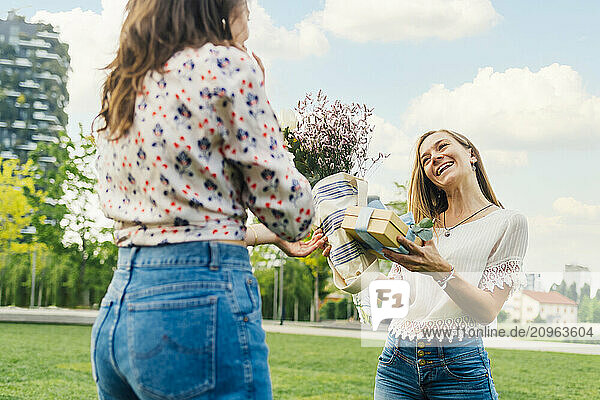 Woman giving gift box and bouquet of flowers to happy friend under sky