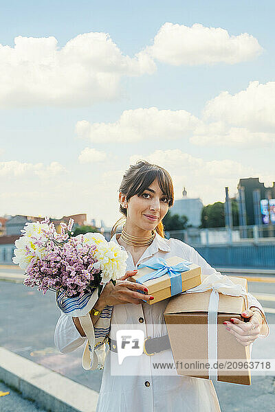 Smiling woman standing with gift boxes and bunch of flowers under cloudy sky