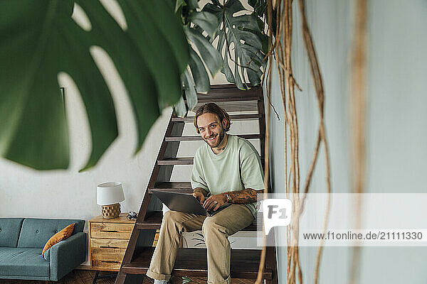 Smiling young freelancer sitting with laptop on staircase at home