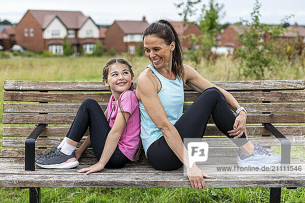 Happy woman sitting back to back with daughter on bench