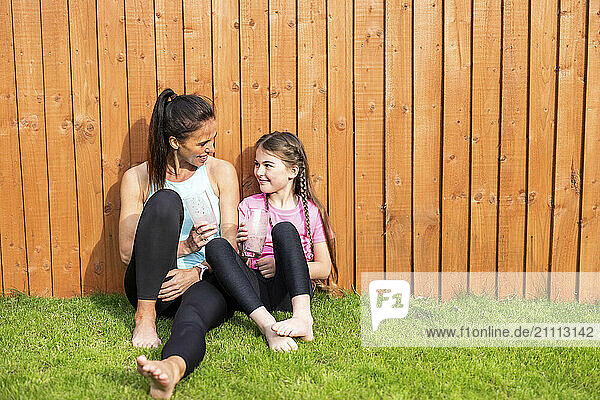 Mother and daughter holding smoothie glass and sitting near wooden fence
