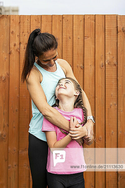 Mother embracing daughter standing in front of wooden fence