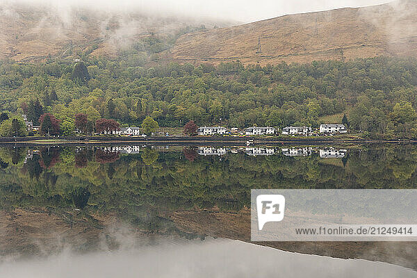 View of a loch with reflections in Glencoe  Scotland  showcasing a serene landscape with houses  hills  and fog.