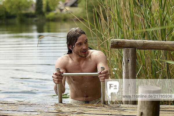 Smiling man emerging from a refreshing swim in a tranquil lake holding onto a wooden dock surrounded by green reeds.