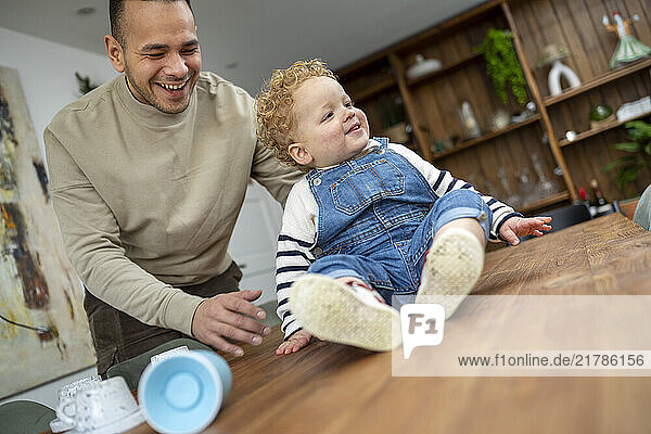 Smiling father with a young child in overalls  sitting on a wooden countertop in a home setting  Netherlands