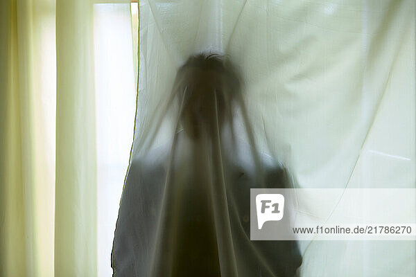 A blurred silhouette of a person behind white curtains exhibiting an abstract and ethereal appearance.