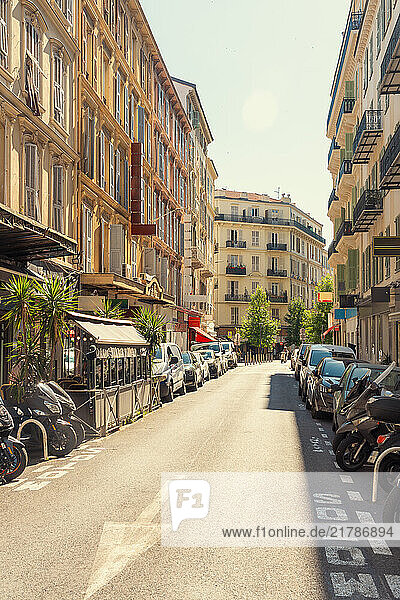 Sunny city street with outdoor cafe seating  lined with traditional European architecture and parked vehicles under a clear blue sky in Nice  France