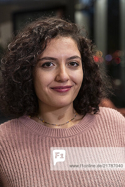 Smiling woman with curly brown hair wearing a pink sweater or pullover indoors  Berlin  Germany